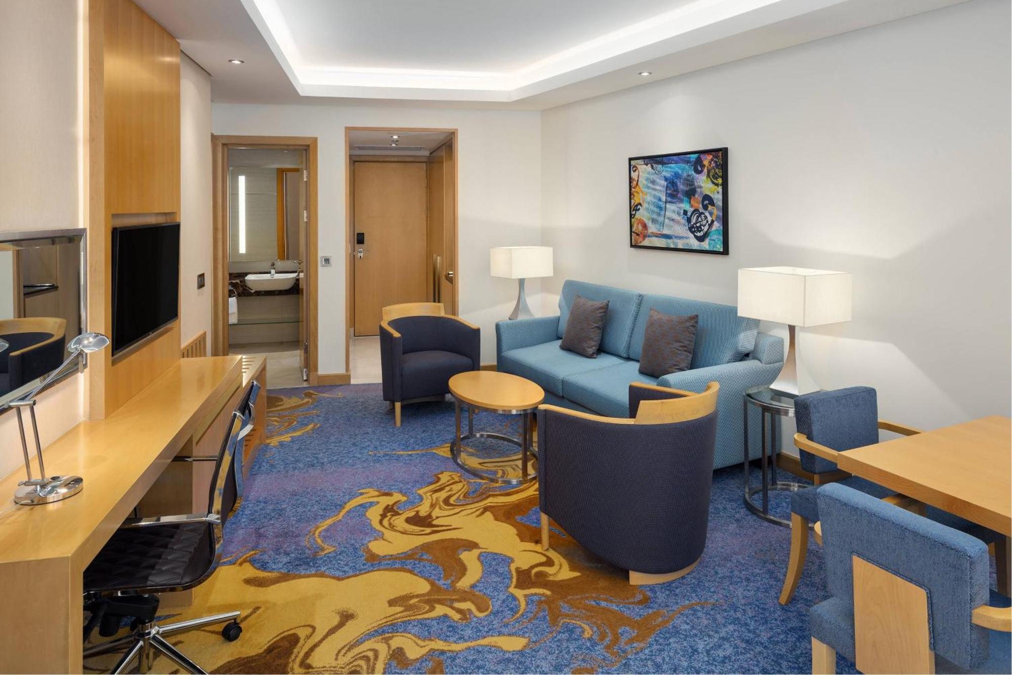 Sheraton Makkah Jabal Al Kaaba Hotel Mekka Exterior foto The photo displays a modern hotel room or suite. The room features a comfortable seating area with a blue couch and two blue armchairs surrounding a small round table. A wooden desk with a black office chair is positioned against one wall, accompanie