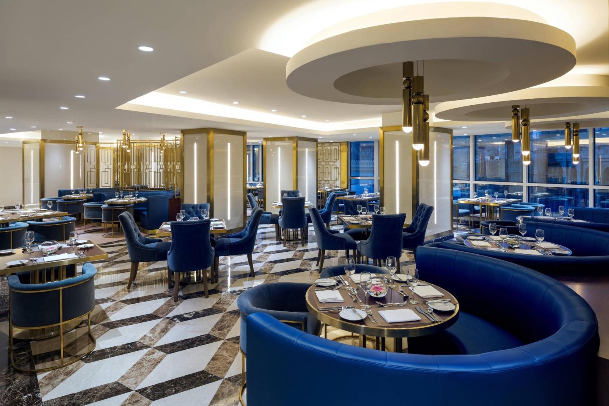 Sheraton Makkah Jabal Al Kaaba Hotel Mekka Exterior foto The photo shows the interior of a modern restaurant. The design features elegant curved blue seating arranged around round tables with plates and glassware set for dining. The flooring is a striking marble pattern, adding to the upscale ambiance. The
